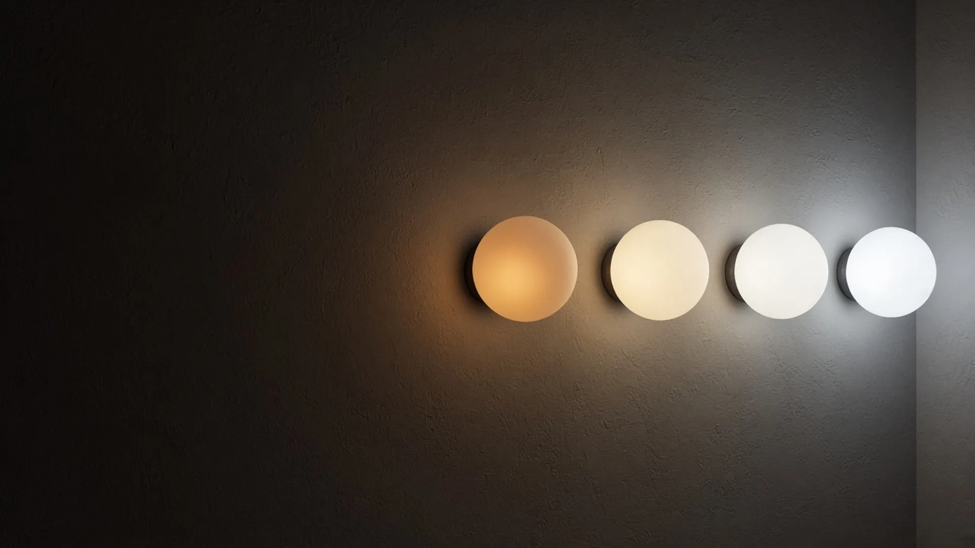 A row of four identical frosted globes glowing from warm amber to cool daylight white against a dark plaster wall