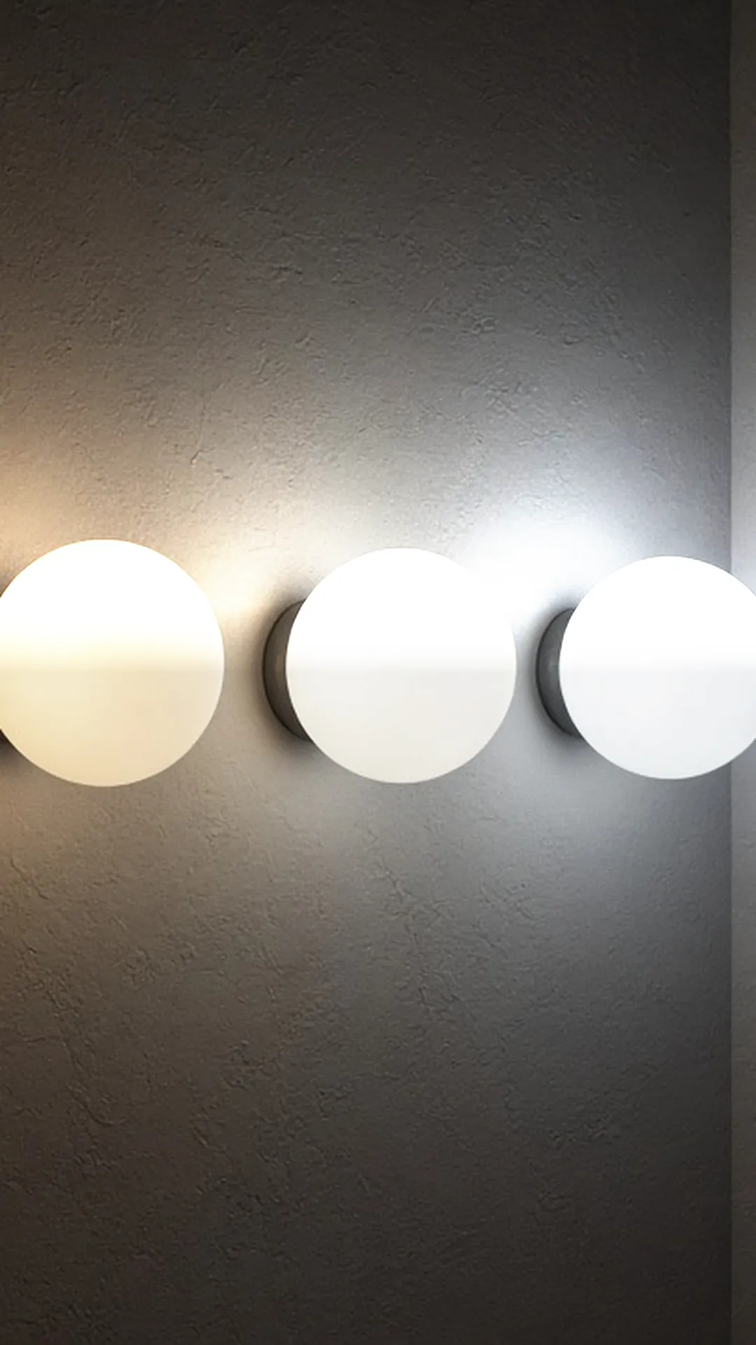 A row of four identical frosted globes glowing from warm amber to cool daylight white against a dark plaster wall