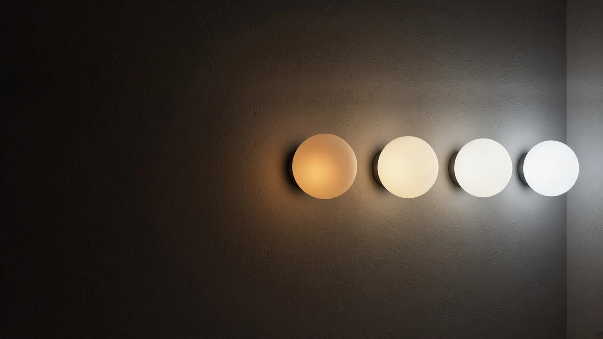 A row of four identical frosted globes glowing from warm amber to cool daylight white against a dark plaster wall